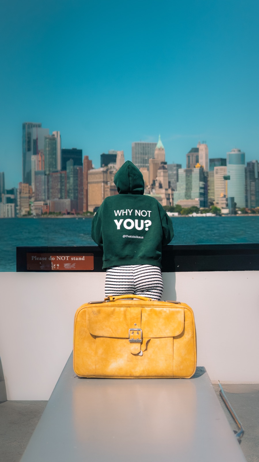 The Why Not You? hoodie looking out at the New York skyline beside the yellow suitcase