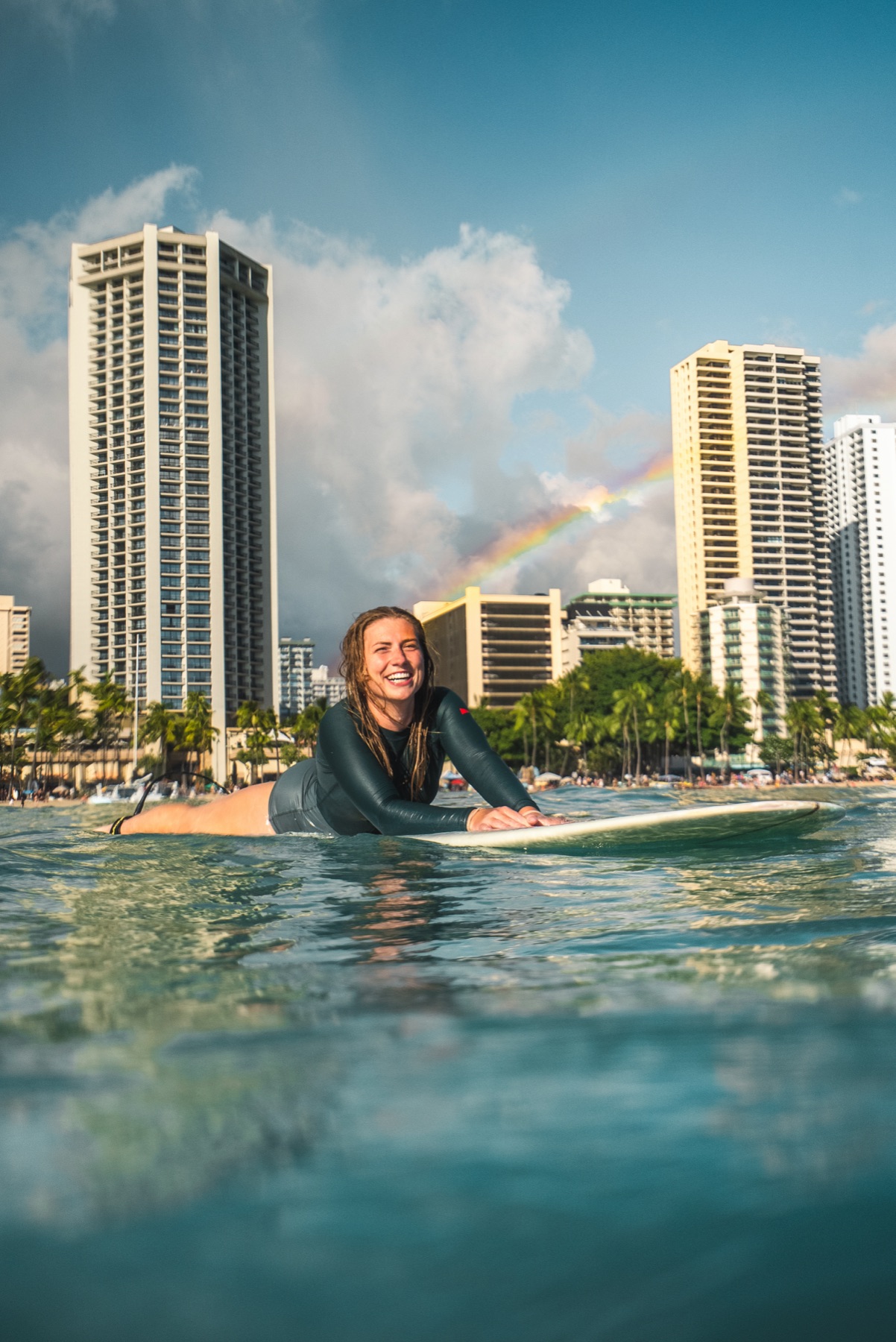 Syd Meyer laughing on her surfboard with the Waikiki hotels behind her